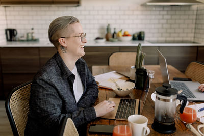 Smiling lesbian professional sitting with laptop at dining table in kitchen