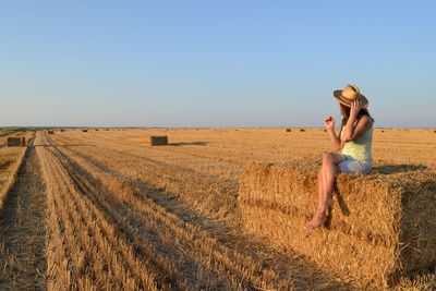 Full length of woman in hat sitting with legs crossed on hay bale against clear sky