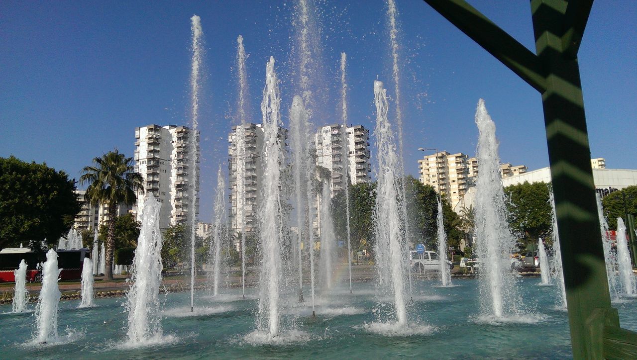 water, fountain, waterfront, tree, clear sky, spraying, motion, splashing, built structure, building exterior, blue, architecture, reflection, nature, day, outdoors, city, sky, sunlight, growth