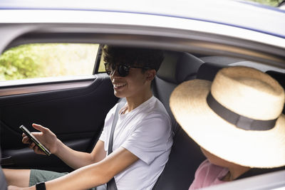 Smiling teenage boy with smart phone wearing sunglasses sitting by brother in car