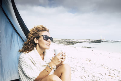 Smiling woman holding drink while sitting at beach by tent against sky