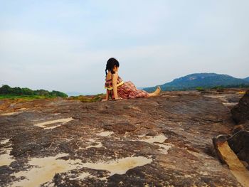 Rear view of girl sitting on rock against sky