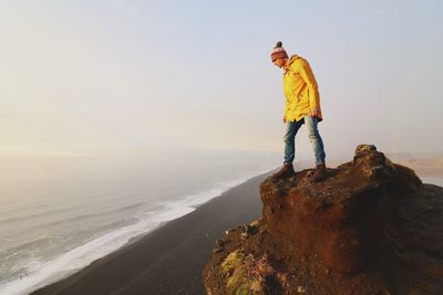 Man standing on rock formation at beach against sky