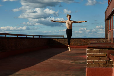 Masterful young blond ballet dancer in black leotard performing exciting jump with raising hands up on roof of building on sky background in sunny day