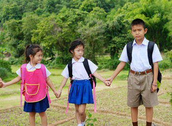 Siblings in school uniforms holding hands while standing against trees