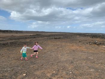 Full length of siblings walking on landscape against cloudy sky