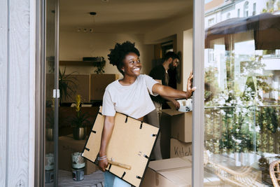 Smiling woman looking away holding frame while leaning on wall at new home