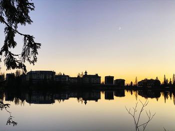 Silhouette buildings by lake against sky at sunset