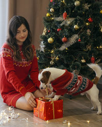 Full length of woman sitting with dog by christmas tree at home