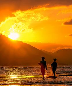 Fishermen walking in shallow water at shore against sky during sunset