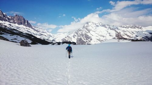 Rear view of hiker walking on snow covered field by rocky mountains against sky