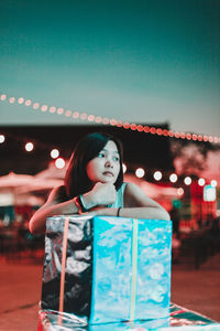 Thoughtful woman leaning on gift box at night