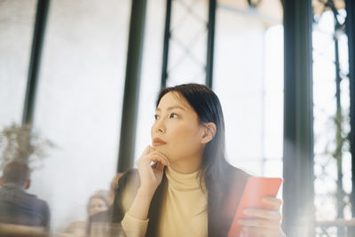 Thoughtful businesswoman holding smart phone while looking away at cafeteria in office