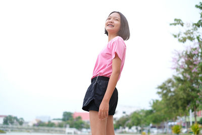 Low angle view of smiling teenage girl looking away while standing against clear sky