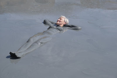 Senior woman with short gray hair relaxing in natural mud bath, fully covered in therapeutic mud