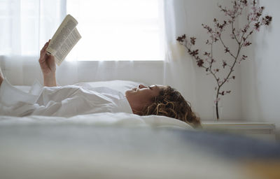 Curly-haired woman reading a book lying on bed.