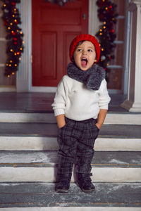 Boy in a sweater and a hat stands on the porch and keeps his hands in his pockets at christmas