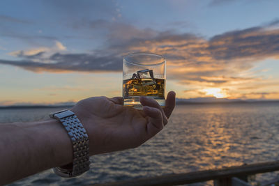 Cropped hand of man holding whisky glass against sea and cloudy sky during sunset