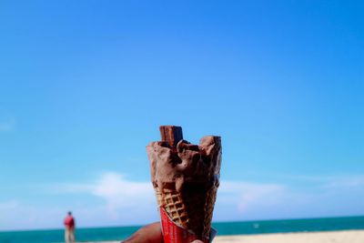 Cropped hand holding ice cream at beach against clear blue sky