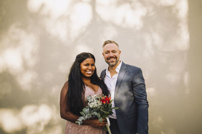 Portrait of smiling bride holding bouquet standing by groom against wall