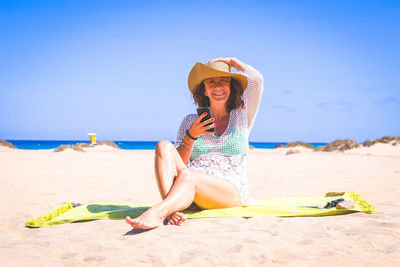 Portrait of smiling mature woman using smart phone while sitting at beach against sky during sunny day