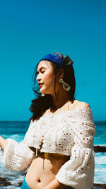 Beautiful young woman standing by sea against blue sky