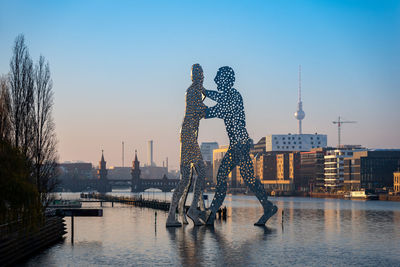 Modern sculpture by river and buildings against clear sky