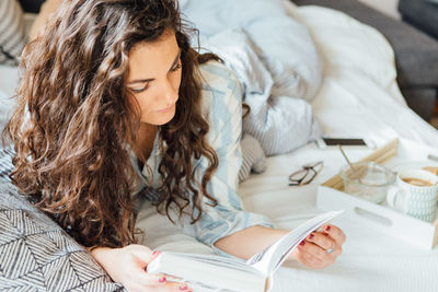 High angle view of young woman reading book while lying on bed at home