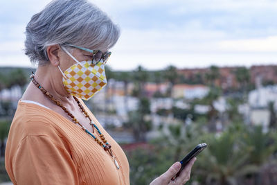 Side view of woman wearing mask using mobile phone against townscape