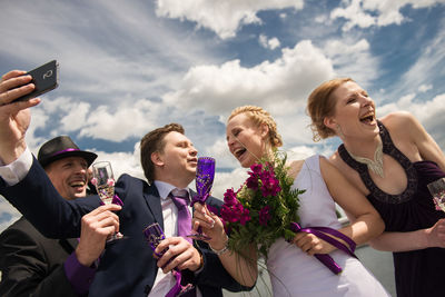 Bridesmaid and best man with bridal couple taking selfie against sky