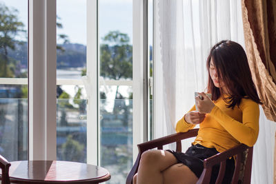 Young woman having drink in cup while sitting on chair at home