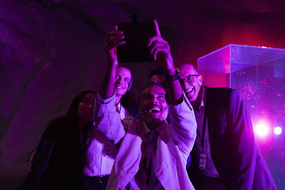 Cheerful businessman taking selfie with colleagues through smart phone at exhibition center