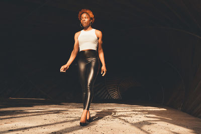 Full body of confident african american female in trendy outfit and high heels standing in dark tunnel with bright sunlight