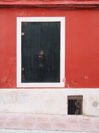 Old dark green wooden closed shutters in a white framed window in an old distressed red painted wall