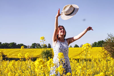 Happy young woman playing with hat while standing amidst yellow flowers on field against sky