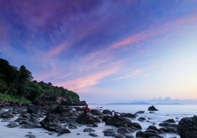 Man sitting on rock at beach against sky during sunset