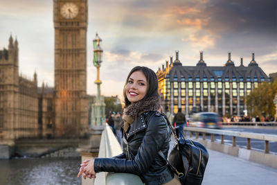 Portrait of smiling young woman standing against tower bridge in city during sunset