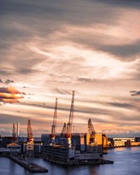 Cranes at commercial dock by sea against sky during sunset