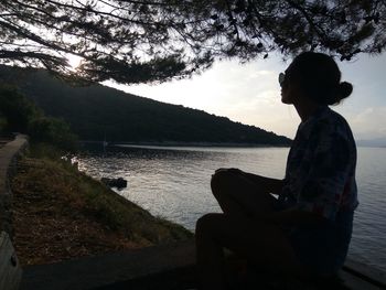 Side view of woman sitting by lake against sky