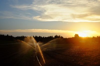 Sprinklers spraying water on field against sky during sunset