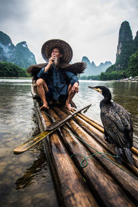 Full length of fisherman with cormorant on wooden raft in river against sky