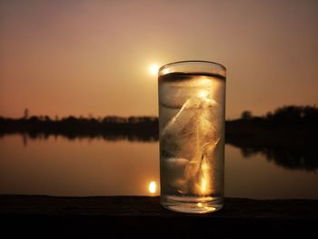 Close-up of glass on table against lake during sunset