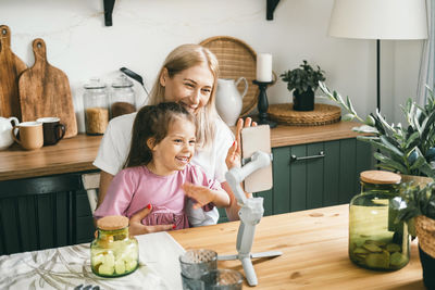 Happy mother and daughter waving hands while looking at webcam in smartphone on tripod for videocall