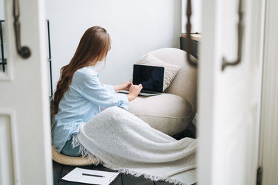 Young smiling woman in blue shirt using laptop in room, view from open doors