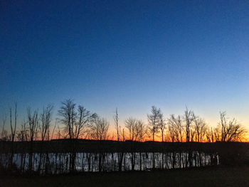 Silhouette bare trees by lake against clear sky during sunset