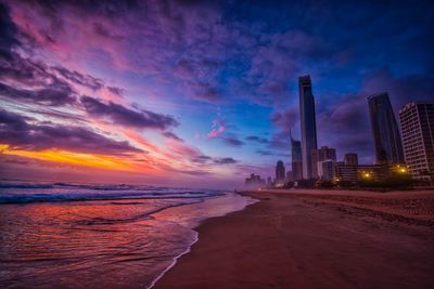 Beach by buildings against sky during sunset