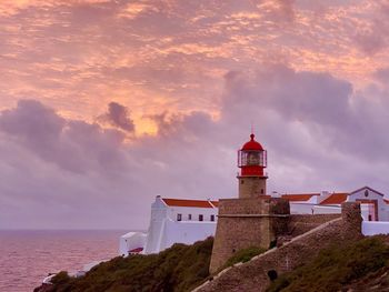 Lighthouse amidst buildings and sea against sky during sunset