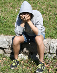 Full length of teenage boy with hands covering face sitting on stones