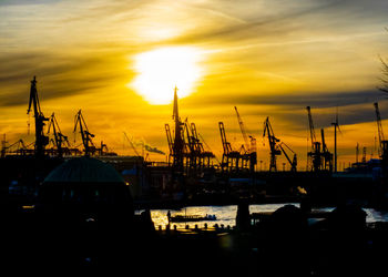 Silhouette cranes at harbor against sky during sunset