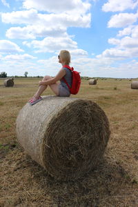 Side view full length of woman sitting on hay bale at agricultural field against sky